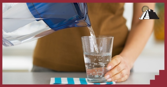 A person pouring water from a pitcher into a clear glass on a counter